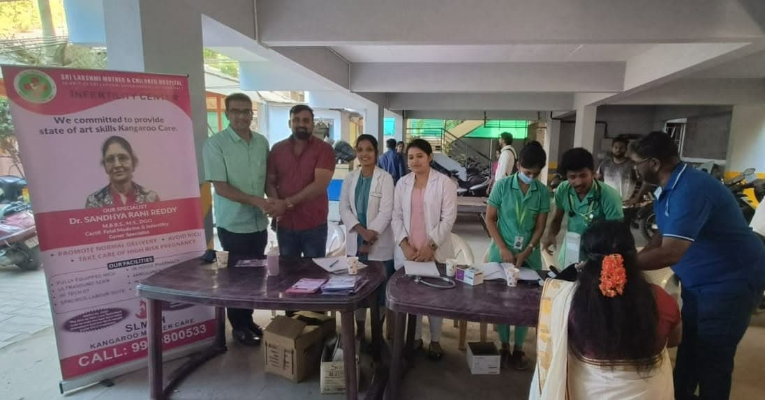 Staff and visitors at a Sri Lakshmi Mother & Children Hospital health camp booth, with an Infertility Center and Kangaroo Mother Care banner in the background.