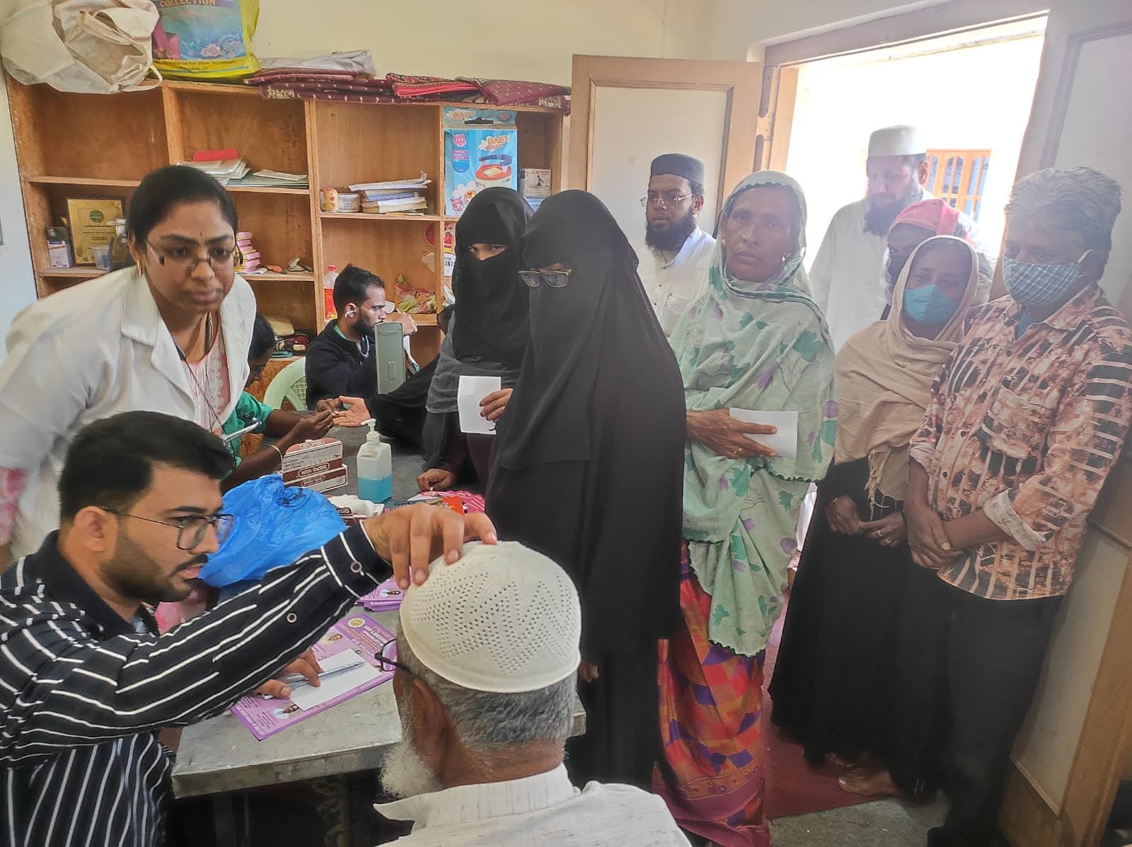 A doctor and hospital staff at a registration desk attending to a group of visitors, including women in burqas and an elderly man, at an indoor free health camp.