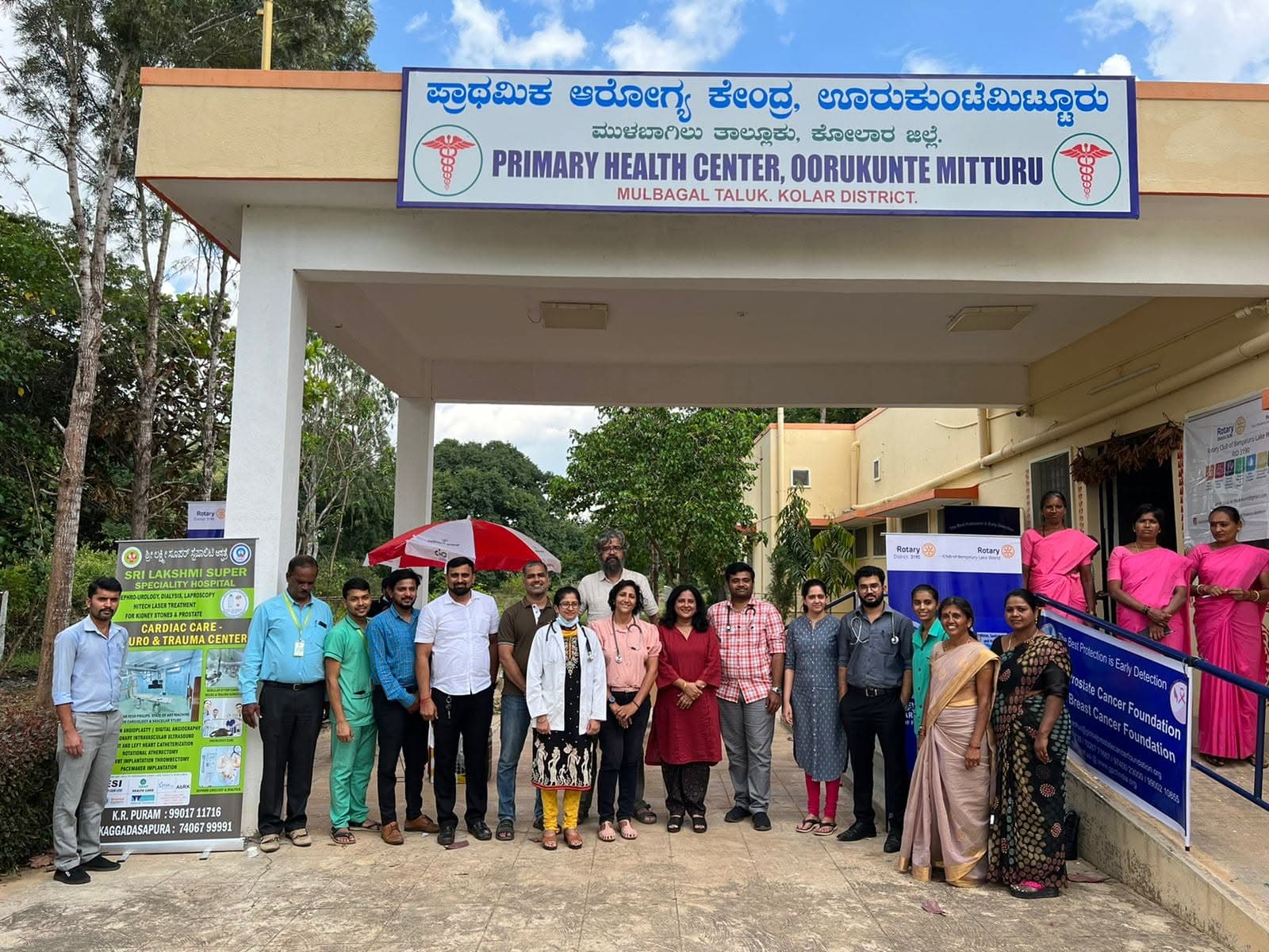 Hospital staff and dignitaries posing outside the Primary Health Center, Oorukunte Mitturu, Mulbagal Taluk, Kolar District, alongside Sri Lakshmi Super Speciality Hospital and Rotary Club banners.