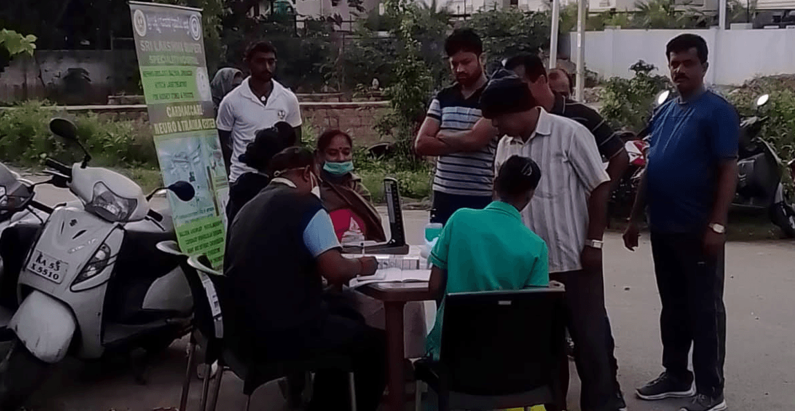 Hospital staff checking a visitor’s blood pressure at an outdoor free health camp table, with a Sri Lakshmi Super Speciality Hospital banner and parked scooters nearby.